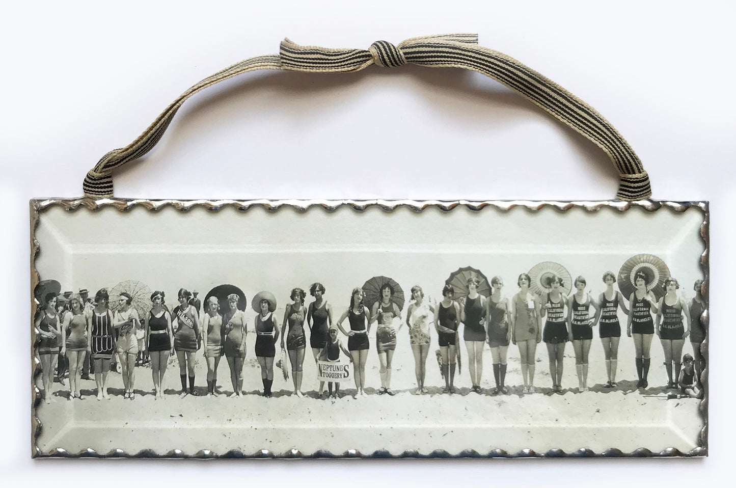 A decorative wall ornament featuring a black and white vintage photograph of women on the beach with umbrellas, with a silver hand soldered frame and beveled glass front.