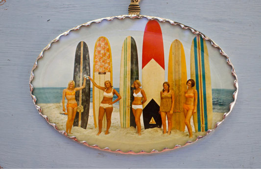 Decorative wall ornament with a vintage-style illustration of women on a beach with surfboards. With silver han soldered frme, and beveled glass front.