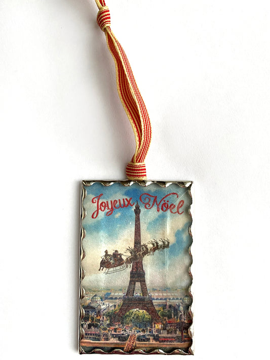 A rectangular ornament with a beveled glass front featuring an illustration of Santa Claus flying over the Eiffel Tower, covered with clear glitter. The ornament has a silver hand soldered frame and a vintage style striped ribbon at the top for hanging.